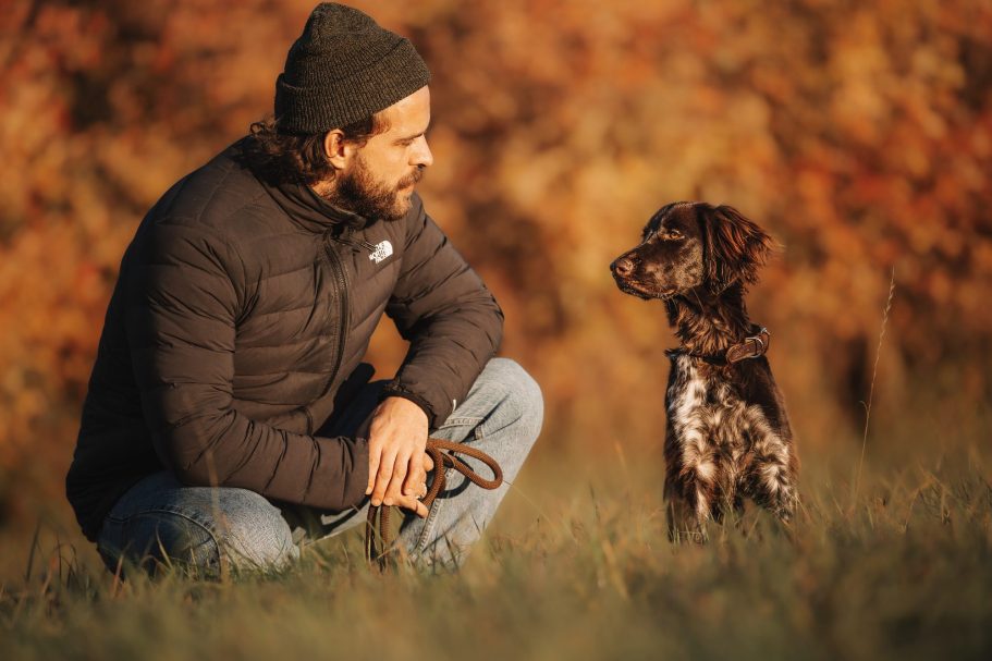 Hundefotografie Junger Mann sitzt auf dem Gras und schaut zu seinem Hund in einer herbstlichen Landschaft. Beziehung Mesch und Tier